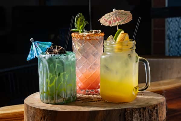 Three colorful tropical cocktails garnished with fruit, herbs, and umbrellas on a wooden table.