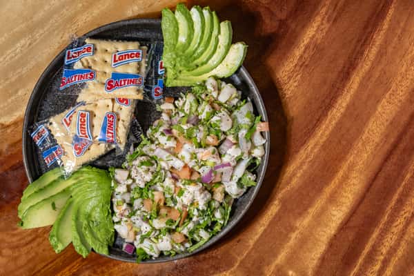 Fresh ceviche topped with cilantro and onion, served with sliced avocado and packets of Lance Saltines on a dark plate.
