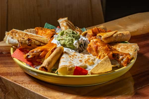 This dish is a festive appetizer platter featuring a mix of Mexican favorites — crispy taquitos, cheesy quesadilla wedges, and saucy buffalo wings — all arranged around a center of creamy guacamole and sour cream, served over colorful tortilla chips for a vibrant presentation.