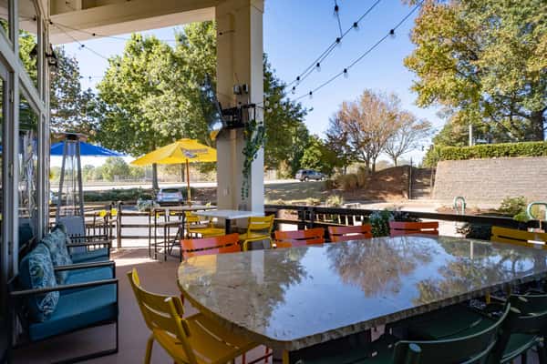 Outdoor patio with colorful chairs and tables, string lights, and large yellow and blue umbrellas under a clear blue sky.