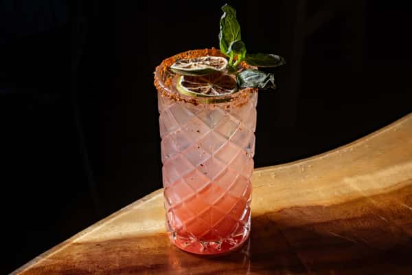 Tall pink cocktail in a textured glass with chili rim, garnished with dried lime slices and fresh basil leaves.
