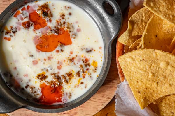 Bowl of queso dip with chorizo and chili oil served alongside crispy tortilla chips on a wooden table.