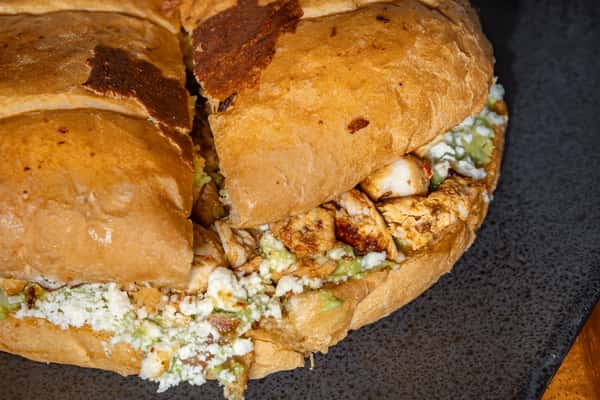 Close-up of a Mexican torta sandwich filled with grilled chicken, crumbled cheese, and guacamole on toasted bread.