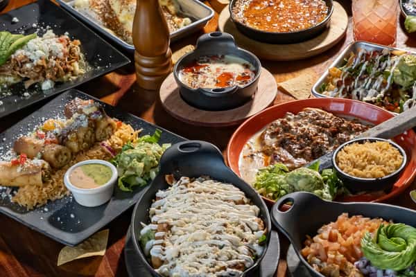 Assorted Mexican dishes on a wooden table, including enchiladas, queso dip, rice, guacamole, and tacos with colorful garnishes.