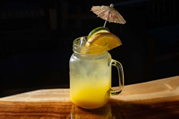 Bright yellow cocktail served in a mason jar with lime, orange slices, and a paper umbrella garnish on a wooden bar.