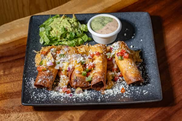 Plate of crispy taquitos topped with diced peppers and cheese, served with guacamole, rice, and a side of creamy dipping sauce.