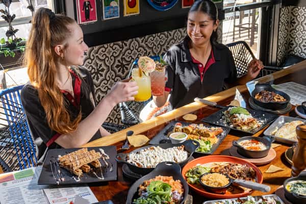 Two women smiling and toasting colorful cocktails over a table filled with vibrant Mexican dishes in a festive restaurant setting.