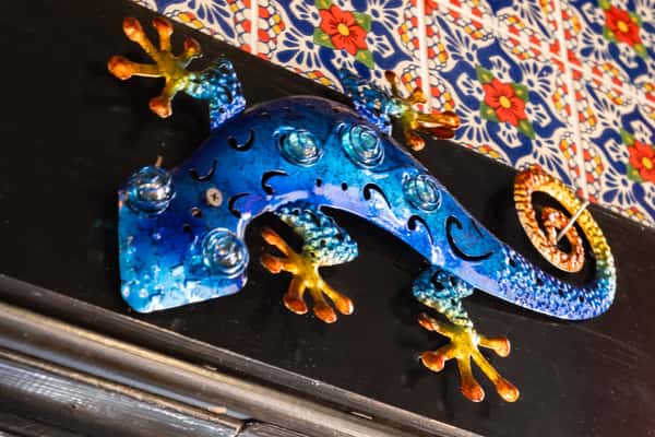 Colorful metal gecko wall art in blue, yellow, and orange tones against a vibrant floral tile background.