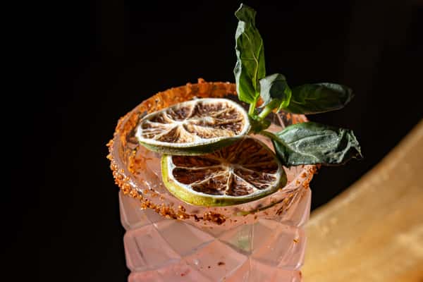 This is a close-up of a vibrant cocktail garnished with dried lime slices, fresh basil leaves, and a chili-salt rim. The drink has a refreshing pink hue, beautifully presented in a textured glass—perfect for a tropical or Mexican-inspired bar setting.