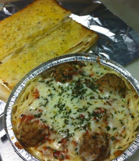 Open garlic bread toasted next to a to-go container of meatballs, tomato sauce, and melted mozzarella on top from different angle