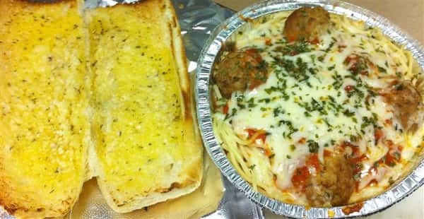 Open garlic bread toasted next to a to-go container of meatballs, tomato sauce, and melted mozzarella on top from different angle