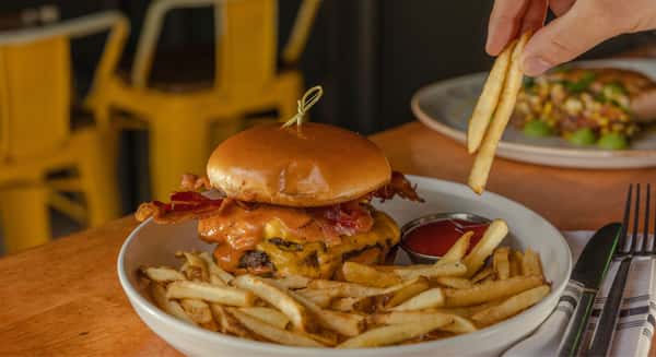 Cheeseburger topped with bacon and melted cheese served with fries as a hand reaches to grab a fry.
