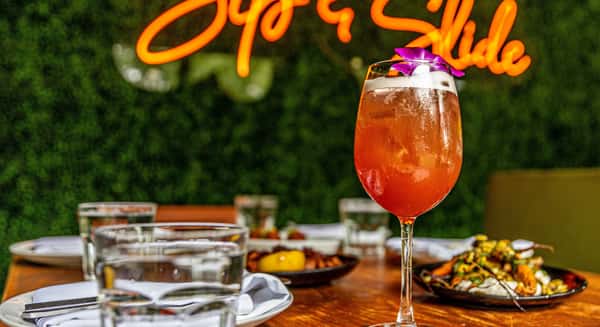Colorful tropical cocktail with orchid garnish on outdoor restaurant table in Coconut Grove, glowing "Sip & Slide" neon sign in background.