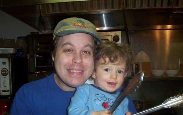 a chef holding a toddler in the kitchen - the toddler is holding cooking tongs