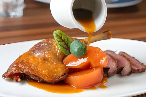 Sliced roasted meat with carrots on a plate while sauce is being poured from a small pitcher.