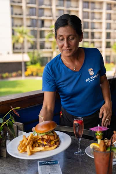 restaurant staff member serving a plated burger and fries at an outdoor coastal dining table