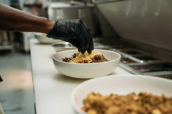 Chef garnishing a hearty bowl of seasoned beef and shredded cheese in a commercial kitchen, showcasing fresh, made-to-order comfort food.
