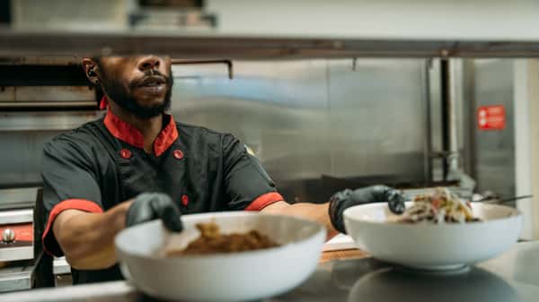 Chef in black and red uniform plating freshly prepared dishes in a commercial kitchen, ready for service.