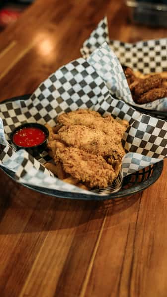 Crispy golden fried chicken tenders served with seasoned fries and a side of tangy red dipping sauce, presented in a checkered basket for a classic comfort food experience.