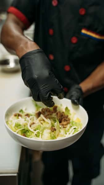 Chef garnishing a fresh taco salad with seasoned beef, shredded cheese, and crisp lettuce in a white bowl, prepared in a professional kitchen.