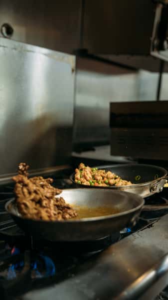 Sizzling pans on a commercial stove cooking seasoned meats and vegetables, showcasing fresh ingredients being prepared in a professional kitchen.