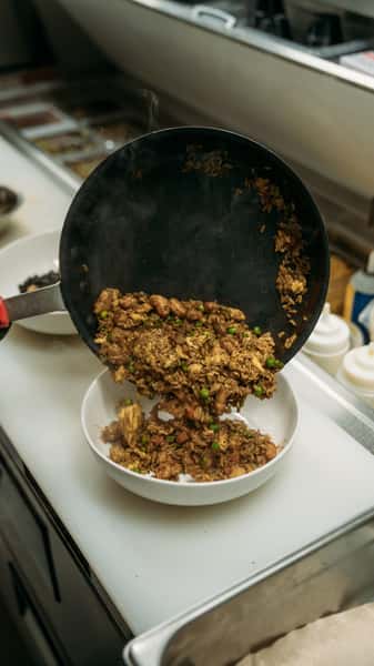 Close-up of hot fried rice with tender chicken, peas, and savory spices being poured fresh from a skillet into a bowl, ready to serve.