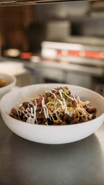 Freshly prepared burrito bowl with seasoned beef, shredded cheese, pico de gallo, and a drizzle of sour cream, served in a white bowl at a restaurant kitchen counter.