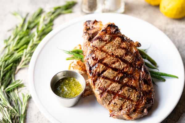 Savor the succulent flavors of our hand-cut 14oz Ribeye Steak at Black+Haus Tavern in Littleton, Colorado & Parker, Colorado. This hearty dish features tender ribeye grilled to perfection, accompanied by roasted potatoes drizzled with garlic aioli and crisp green beans. Topped with zesty chimichurri sauce, our gluten-free ribeye steak celebrates local ingredients and gourmet cuisine. Whether you're planning a romantic dinner, celebrating a special occasion, or enjoying a family-friendly dining experience, our 14oz Ribeye Steak promises a memorable culinary journey at Littleton's favorite restaurant. Explore our seasonal menu and indulge in the best flavors of Black+Haus Tavern.