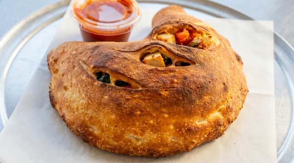 Calzone with a crispy golden-brown crust, revealing chicken, spinach, and tomatoes inside, served with a side of marinara sauce.