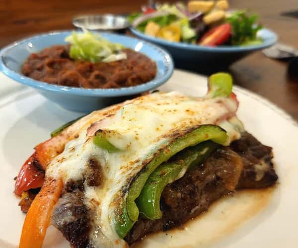 Grilled steak topped with melted cheese and sautéed bell peppers, served with seasoned beans and a side salad.
