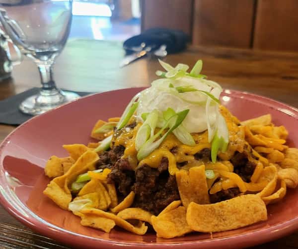 Frito pie topped with seasoned ground beef, melted cheddar cheese, sour cream, and sliced green onions in a red bowl.