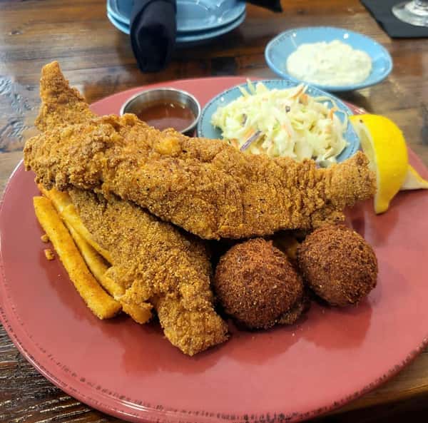 Southern-style fried catfish served with fries, hush puppies, coleslaw, tartar sauce, and a lemon wedge.
