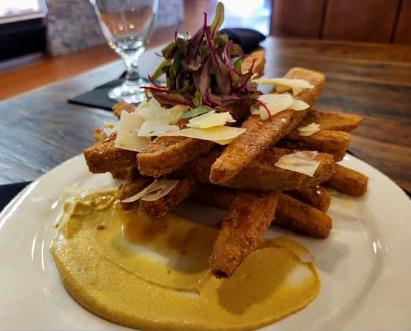 Crispy fried polenta fries stacked neatly, topped with shaved Parmesan and microgreens, served with a generous smear of creamy mustard dipping sauce.