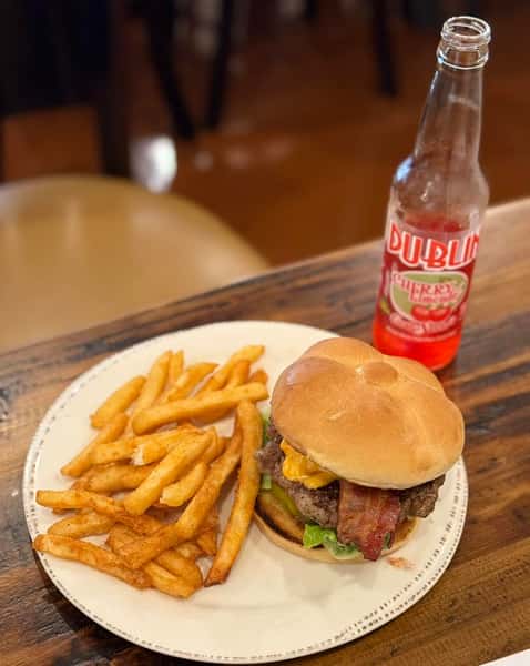 Cheeseburger with bacon, lettuce, and fries on a white plate, paired with a bottle of cherry soda on a wooden table.