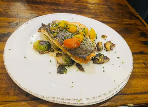 This dish appears to be a beautifully seared fish fillet—likely sea bass or trout—topped with cherry tomatoes, capers, and a light herb garnish. It's served alongside roasted Brussels sprouts. The crispy skin and fresh, colorful toppings suggest a Mediterranean-style preparation.