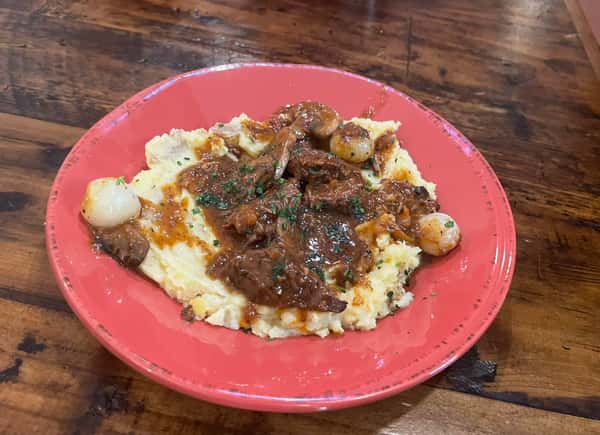 This dish appears to be a classic beef stew or pot roast served over creamy mashed potatoes. It's topped with a rich brown gravy and garnished with pearl onions and chopped herbs—comfort food at its finest.