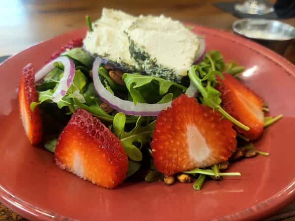 Fresh arugula salad topped with sliced strawberries, red onion, spiced pecans, and herbed goat cheese on a red plate.