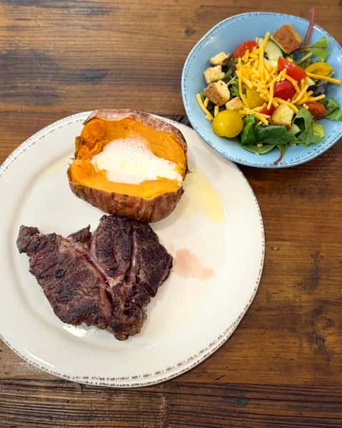 This plate features a classic hearty meal: a grilled T-bone steak paired with a baked sweet potato topped with melting butter and a drizzle of syrup or honey. It's accompanied by a fresh garden salad in a blue bowl, complete with shredded cheddar cheese, cherry tomatoes, cucumbers, and croutons. A wholesome, comforting combination.