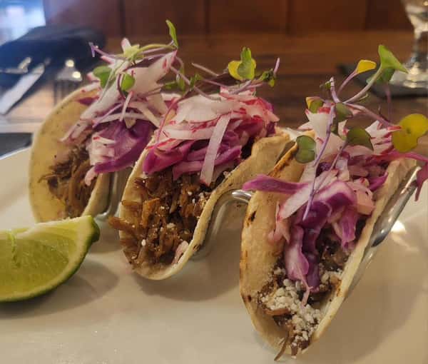 Three shredded meat street tacos topped with purple cabbage slaw, radish, and microgreens, served with a lime wedge.
