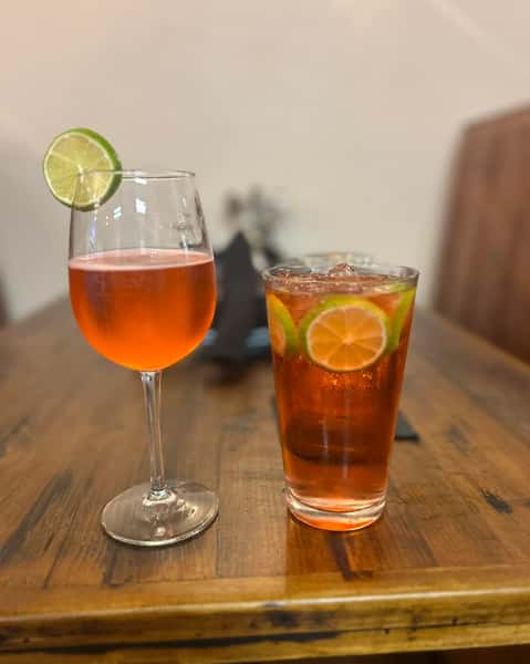 Two vibrant orange cocktails, one in a wine glass with lime garnish and the other in a tall glass with lime slices and ice.
