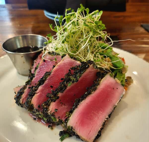 Sesame-crusted seared ahi tuna slices served with sprouts, avocado salad, and a side of soy-based dipping sauce.