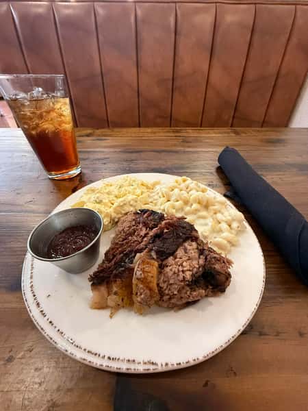 Barbecue brisket with a side of macaroni and cheese, coleslaw, and barbecue sauce, served with iced tea on a wooden table.