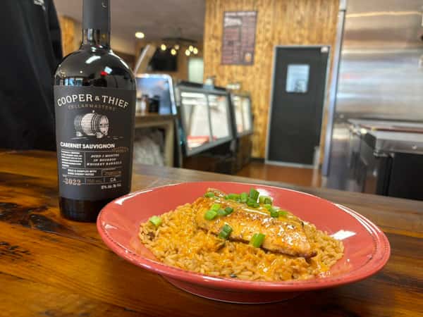 Grilled fish fillet over seasoned rice with creamy sauce and scallions, served beside a bottle of Cooper & Thief Cabernet Sauvignon.