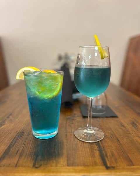 Two blue cocktails on a wooden table, each garnished with citrus slices, one served in a highball glass and the other in a wine glass.