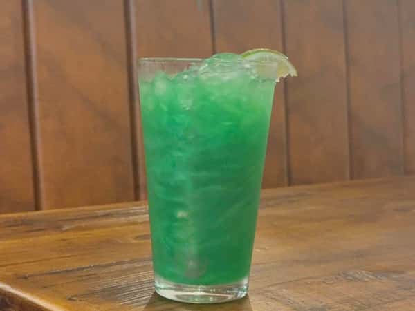 Bright green cocktail served over crushed ice in a tall glass, garnished with a lime wedge on a wooden table.
