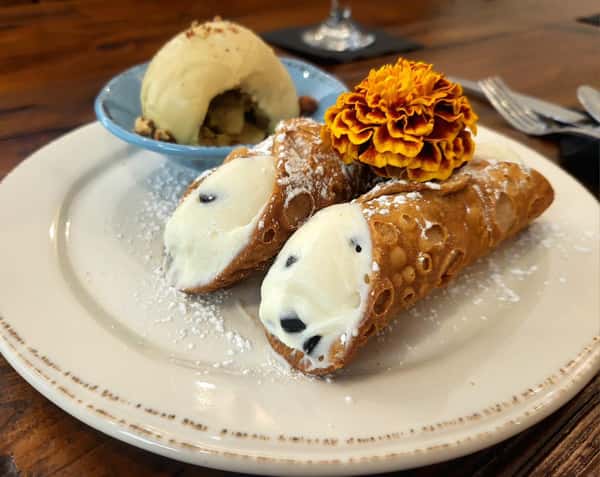 Two chocolate chip cannoli dusted with powdered sugar, served with a scoop of vanilla ice cream and a marigold flower garnish.