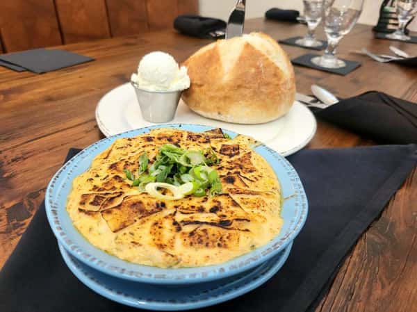 Cheesy baked dip topped with green onions, served in a blue bowl alongside a round loaf of bread and a scoop of whipped butter.