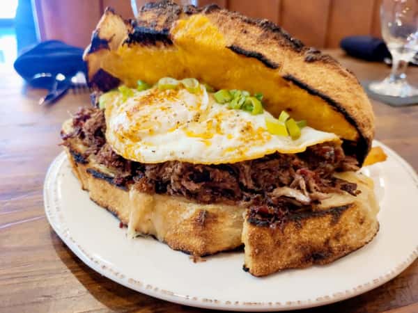 Pulled pork sandwich with melted cheese, topped with a fried egg and green onions, served on toasted rustic bread.
