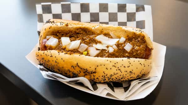 Hot dog in a poppy seed bun with chili and diced white onions, served in a paper tray.