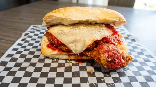 Meatball sandwich with marinara sauce, melted mozzarella, and grated parmesan on a white bun, on checkered paper.
